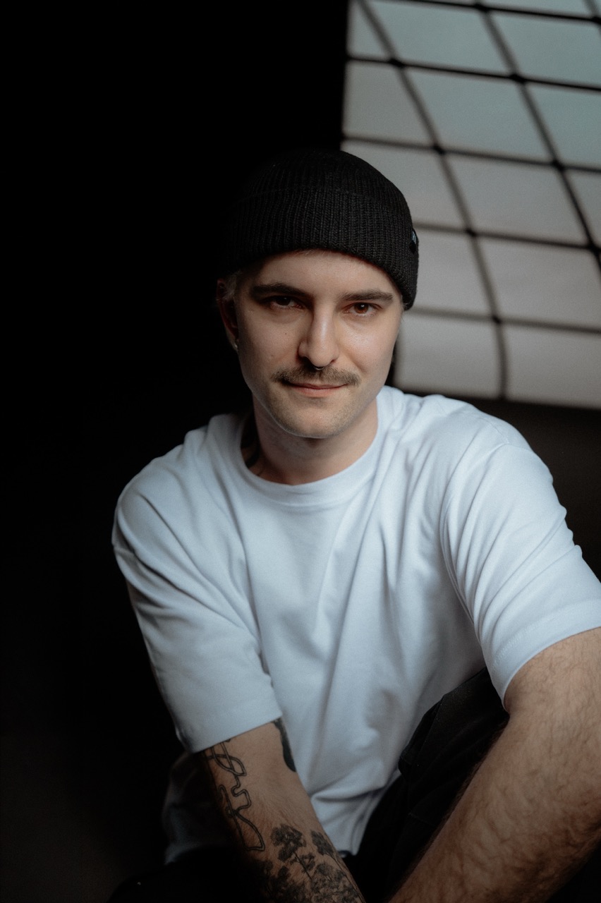 Portrait of Alexander Holzapfel: black beanie, white t-shirt, looking into the camera.